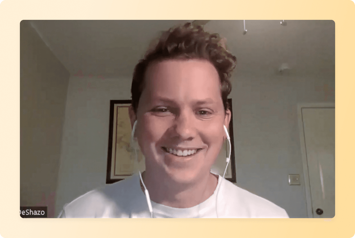 Smiling person with curly hair wearing white t-shirt and earbuds during video call in home office with framed artwork